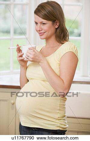 Stock Image - Pregnant woman standing in kitchen with coffee and cigarette smiling. Fotosearch