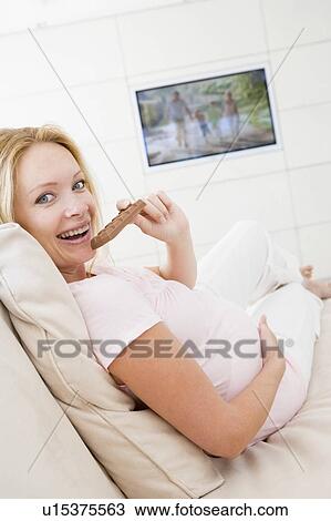 Stock Image - Pregnant woman watching television and eating chocolate smiling. Fotosearch