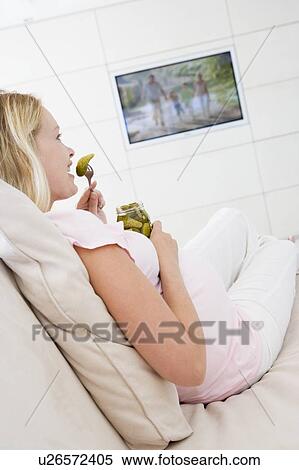 Stock Photography - Pregnant woman watching television and eating pickles smiling. Fotosearch