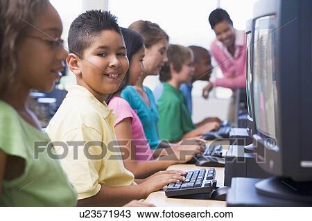 Stock Image - Six children at computer terminals with teacher in background (selective focus/high key). Fotosearch