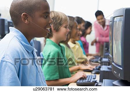 Stock Image - Six children at computer terminals with teacher in background (depth of field/high key). Fotosearch