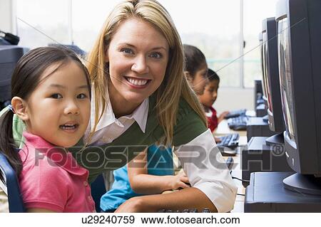 Stock Photo - Teacher helping student at computer terminal with students in background (depth of field/high key). Fotosearch