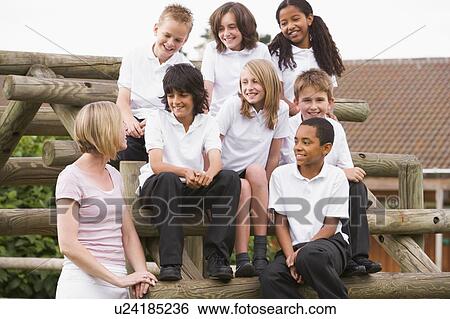 Stock Photograph - Seven students sitting on wooden structure with teacher standing beside them. Fotosearch