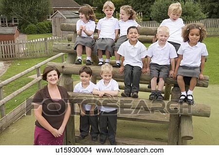 Students sitting outdoors on wooden structure with teacher standing beside them View Large Photo Image Picture - Students sitting outdoors on wooden structure with teacher standing beside them. Fotosearch