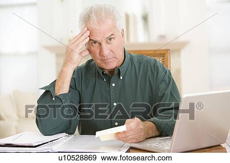 Stock Photo - Man in dining room with laptop and paperwork looking frustrated. Fotosearch