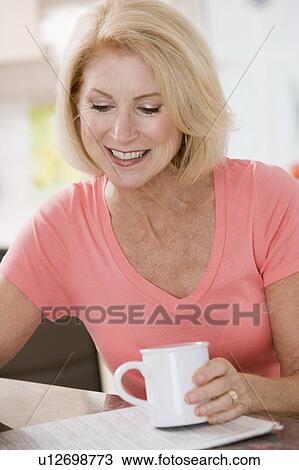 Stock Image - Woman in kitchen with coffee and newspaper smiling. Fotosearch