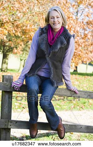 Stock Photo - Woman outdoors at park sitting on fence smiling (selective focus). Fotosearch