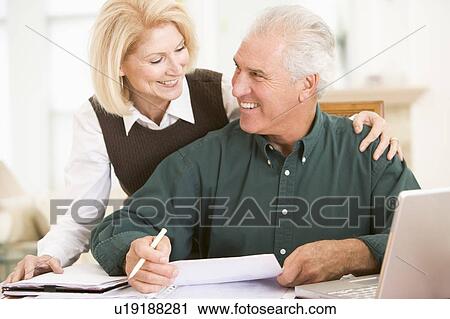 Stock Image - Couple in dining room with laptop and paperwork smiling. Fotosearch