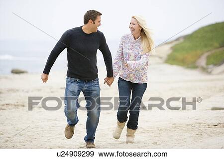 Stock Photo - Couple running at beach holding hands smiling. Fotosearch