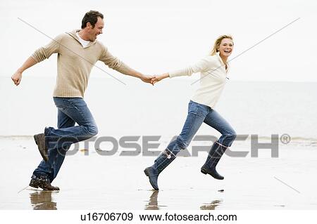 Stock Photo - Couple running on beach holding hands smiling. Fotosearch