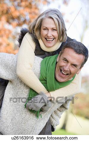 Stock Photograph - Senior man outdoors piggybacking woman and smiling (selective focus). Fotosearch