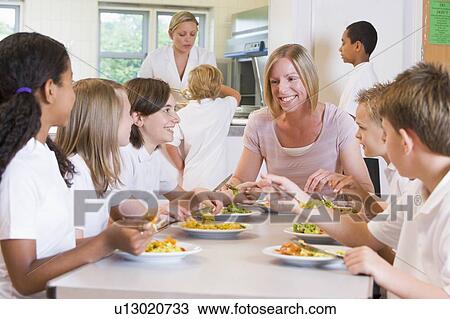 Stock Image - Students sitting at cafeteria table eating lunch with teacher. Fotosearch