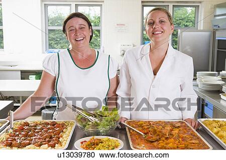 Stock Image - Two lunch ladies standing behind full lunch service station. Fotosearch