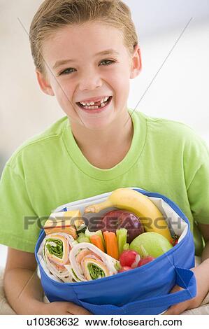 Stock Image - Young boy holding packed lunch in living room smiling. Fotosearch