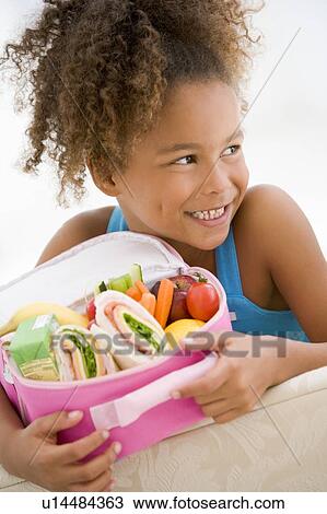 Stock Image - Young girl holding packed lunch in living room smiling. Fotosearch