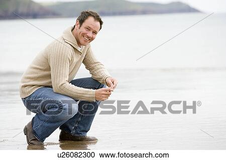 Stock Image - Man crouching on beach smiling. Fotosearch