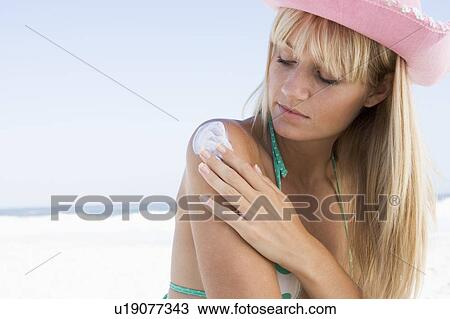 Stock Image - Woman on beach applying sunblock lotion to her shoulder. Fotosearch