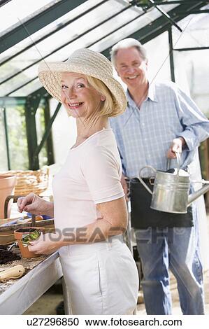 Stock Image - Woman in greenhouse planting seeds and man holding watering can smiling. Fotosearch