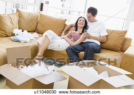 Stock Image - Couple relaxing with champagne by boxes in new home smiling. Fotosearch