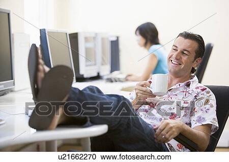 Stock Image - Man in computer room with feet up drinking coffee and smiling. Fotosearch
