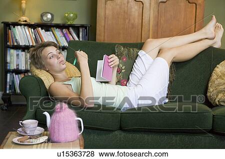 Stock Photo - Young woman lying on sofa at home writing in her diary. Fotosearch