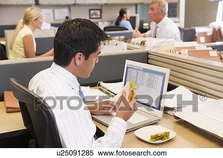 Stock Photography - Businessman in cubicle at laptop eating sandwich. Fotosearch