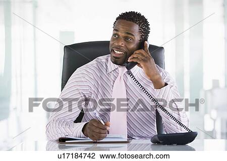 Stock Image - Businessman sitting in office with personal organizer open on telephone. Fotosearch