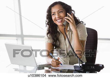 Businesswoman in office on telephone by laptop smiling (high key/selective focus) View Large Photo Image Picture - Businesswoman in office on telephone by laptop smiling (high key/selective focus). Fotosearch