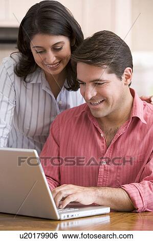 Stock Photograph - Couple in kitchen with paperwork using laptop smiling. Fotosearch