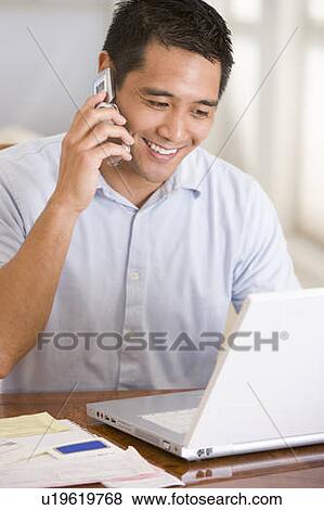 Stock Photo - Man in dining room on cellular phone using laptop smiling. Fotosearch