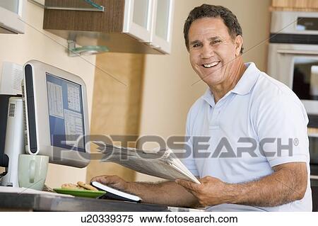 Stock Photography - Man in kitchen with computer and newspaper smiling. Fotosearch