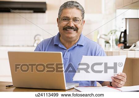 Stock Photo - Man in kitchen with laptop and paperwork smiling. Fotosearch