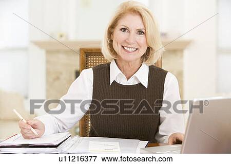 Stock Photo - Woman in dining room with laptop and paperwork smiling. Fotosearch
