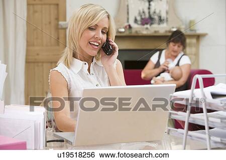 Stock Photograph - Woman in home office with laptop and telephone with mother and baby in background. Fotosearch