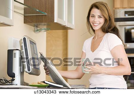 Stock Image - Woman in kitchen at computer with newspaper and coffee smiling. Fotosearch