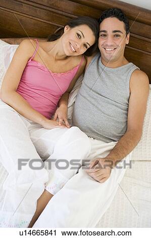 Stock Image - Couple relaxing on bed in bedroom smiling (selective focus). Fotosearch