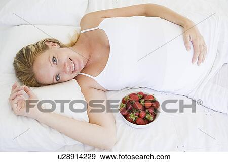 Stock Photo - Pregnant woman lying in bed with bowl of strawberries smiling. Fotosearch