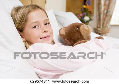 Stock Image - Young Girl Lying In Hospital Bed, Holding Teddy Bear. Fotosearch
