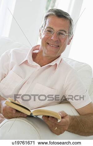 Stock Image - Man in living room reading book smiling. Fotosearch