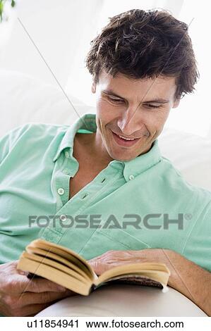 Stock Image - Man reading book in living room smiling. Fotosearch