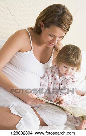 Picture - Pregnant woman in bedroom reading book with daughter. Fotosearch