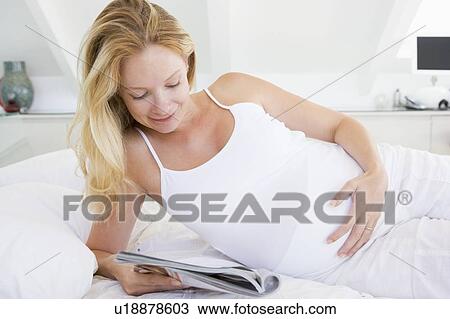 Stock Image - Pregnant woman lying in bed reading magazine smiling. Fotosearch