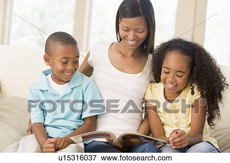 Stock Photo - Woman and two children sitting in living room reading book and smiling. Fotosearch