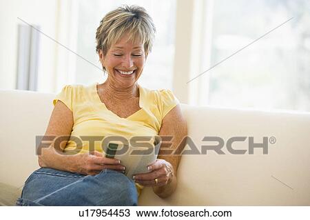 Stock Image - Woman in living room reading book smiling. Fotosearch