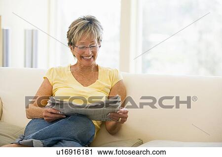 Stock Image - Woman relaxing with newspaper in living room smiling. Fotosearch