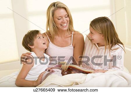 Stock Photo - Woman with two young children sitting in bed reading book and smiling. Fotosearch