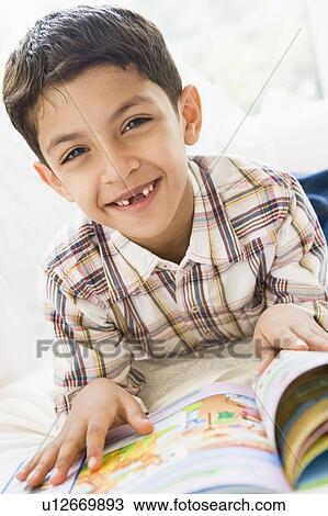 Stock Image - Young boy in living room reading a book and smiling (high key). Fotosearch