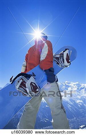 Stock Image - Snowboarder walking on snowy hill carrying board (lens flare). Fotosearch