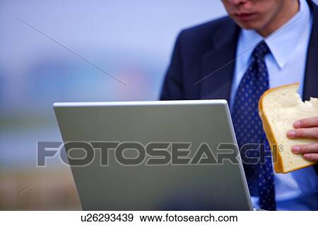Stock Photo - Young businessman eating toast while using laptop. Fotosearch