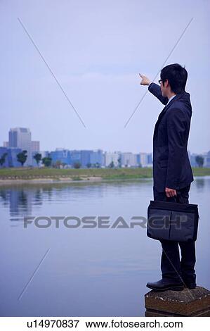 Stock Photo - Young businessman holding briefcase pointing at distance. Fotosearch